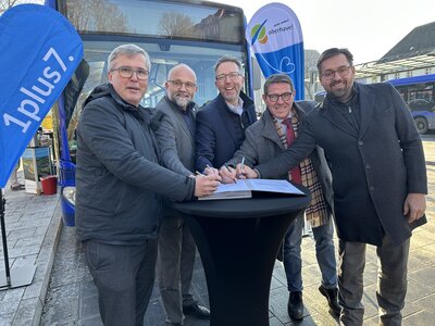 Fünf Männer mittleren Alters stehen um einen schwarzen Stehtisch mit Stiften in den Händen. Im Hintergrund ein Verkehrsbus und zwei Beachflaggen mit dem Logo 1plus7 und vom Landkreis Oberhavel. 