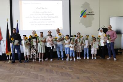 Eine Gruppe von sechs Erwachsenen und acht Kindern steht in einer Reihe vor einer Wand. Alle halten eine Sonnenblume in der Hand. An der Wand sieht man das projizierte Gelöbnis zur Einbürgerung des Landkreises Oberhavel.