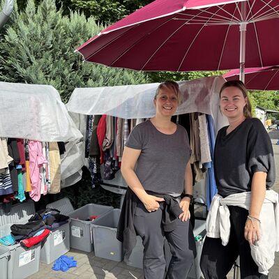 Zwei Frauen stehen vor Kleiderständen und Kisten voller Kleidungsstücke.