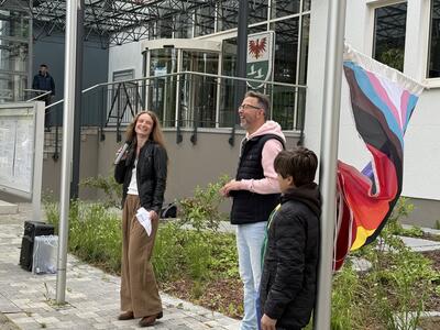 Eine junge Frau und ein Mann mittleren Alters stehen zwischen zwei Fahnenmasten. Ganz rechts im Bild ist ein kleiner Junge mit schwarzer Jacke und dunklen Haaren zu sehen.