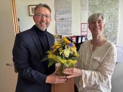 Ein Mann und eine Frau stehen in einem Büro. Sie halten gemeinsam eine Vase mit einem Blumenstrauß in den Händen. 