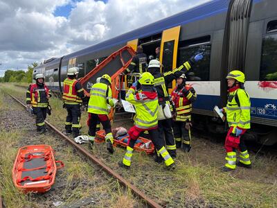 Männer in neongelben Westen mit Schutzhelmen und orangefarbenen Rettungstragen stehen vor einem blau-weißen Zug mit geöffneten, gelben Türen. 