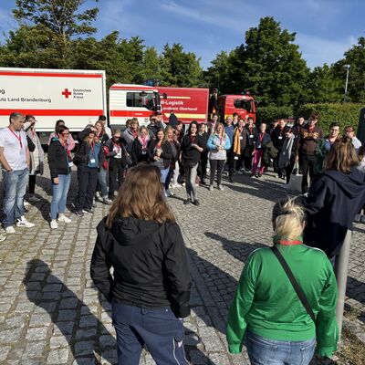 Viele weibliche und männliche Personen stehen in einem Halbkreis. Drumherum Notfall- und einsatzfahrzeuge von Feuerwehr und Katastrophenschutz.