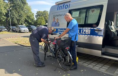 Zwei Männer - der Mann links vorgebeugt in Polizeiuniform - stehen vor einem Polizeieinsatzwagen und untersuchen ein Fahrrad. 