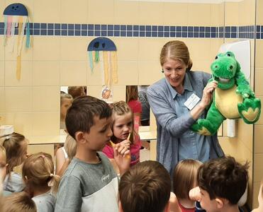 Eine Frau hält ein Krokodil in den Händen. Im Vordergrund putzen sich Kinder im Kitaalter die Zähne. 
