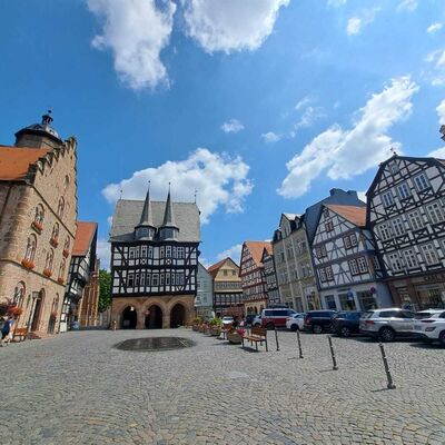 Das Bild zeigt einen Platz mit Fachwerkhäusern und gepflastertem Boden unter blauem Himmel mit vereinzelten Wolken.