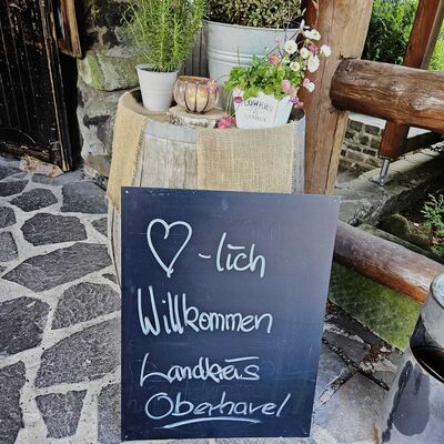 Ein gestaltetes Willkommensschild mit der Aufschrift »Herzlich willkommen Landkreis Oberhavel« steht vor einem Holzfass, dekoriert mit Blumen und Kräutern.