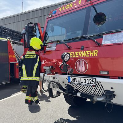 Zu sehen ist ein Feuerwehrfahrzeug. Auf der Linkes Seite des Feuerwehrautos hängt ein kompletter Einsatz-Anzug inklusive Helm.
