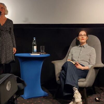 Eine Frau mit kinnlangen, blonden Haaren steht mit einem Mikrofon in der rechten Hand links auf dem Bild neben einem blauen Tisch. Rechts im Bild sitzt eine Frau mit Brille und braunen Haaren in einem hellgrauen Sessel und hat ein Buch auf dem Schoß.