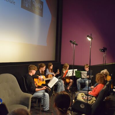 Jungs und Mädchen sitzen mit Musikinstrumenten vor drei kleinen Scheinwerfern. Im Hintergrund ist eine große Beamerleinwand zu sehen. Rechts im Bild sitzen Männer und Frauen im Publikum.