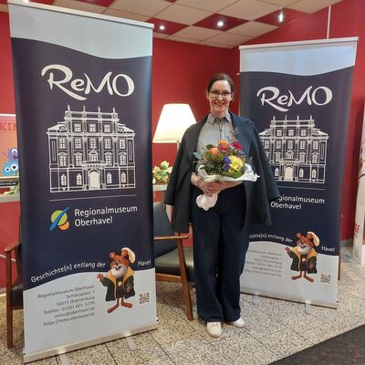 Eine Frau mit Brille und dunkelbraunen, zum Zopf gebundenen Haaren steht mit einem Blumenstrauß in den Händen vor zwei Roll-up Bannern des ReMO - Regionalmuseum Oberhavel. Bei der Frau handelt es sich um die Autorin Alex Beer.