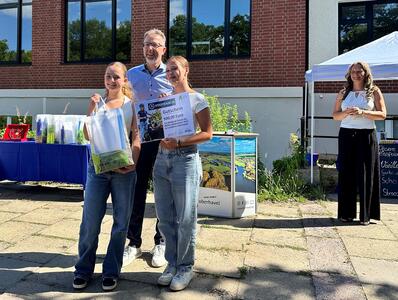 Zwei junge Mädchen halten Plakate in die Höhe. Hinter ihnen steht ein großer Mann mit blauem Hemd in der Mitte. Im Hintergrund eine Frau mittleren Alters mit lockigen, langen braunen und weißem Oberteil und schwarzer Hose. 