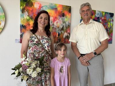 Eine Frau mit schulterlangen braunen Haaren, einem sommerlichen Kleid mit Blumenmuster und einem Blumenstrauß in der Hand steht zusammen mit einem jungen Mädchen und einem älteren Herrn mit grauem Schnäuzer vor einer Wand mit farbenfrohen Bildern. 
