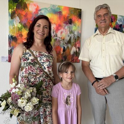 Eine Frau mit schulterlangen braunen Haaren, einem sommerlichen Kleid mit Blumenmuster und einem Blumenstrauß in der Hand steht zusammen mit einem jungen Mädchen und einem älteren Herrn mit grauem Schnäuzer vor einer Wand mit farbenfrohen Bildern.