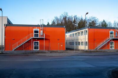 Außenaufnahme. Im Vordergrund ist eine Straße zu sehen, im Hintergrund stehen zwei orangefarbene Containergebäude mit Treppenaufgängen.