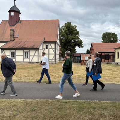 Gruppenfoto von Männern und Frauen auf einem Dorfweg. Im Hintergrund eine alte Dorfkirche.
