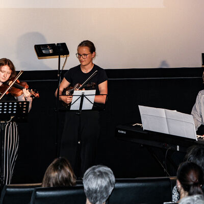 Drei Schülerinnen der Kreismusikschule spielen auf Geigen und einem Keyboard vor dem Publikum. Im Hintergrund befindet sich eine beleuchtete Leinwand.