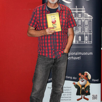 Der Autor Peter Grandl vor dem Roll-up Banner des ReMO - Regionalmuseum Oberhavel nach seiner Lesung am 30.06.2025 während er sein Buch "Reset" hochhält.