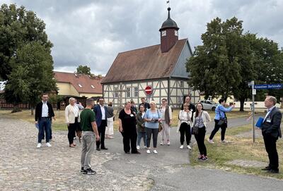 Eine Gruppe von mehreren Männern und Frauen stehen auf der Kreweliner Dorfstraße. Im Hintergrund ist eine alte Dorfkirche zu sehen.
