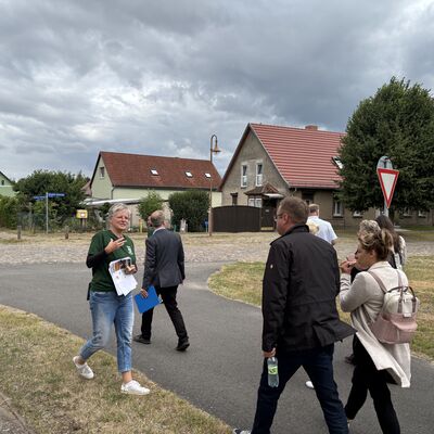 Eine Gruppe von Männern und Frauen läuft auf einem Dorfweg. Im Hintergrund sind einzelne Dorfhäuser zu sehen.
