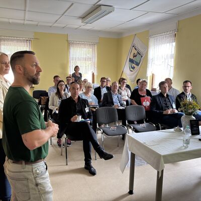 Eine Gruppe von Männern und Frauen sitzt in einem Raum mit gelben Wänden auf Stühlen in Viererreihen. Vorne im Bild ein Mann mit Bart und grünem Shirt und eine blonde Frau mit gelbem Sakko.