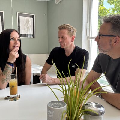 Eine Frau und zwei Männer sitzen an einem weißen Cafétisch und unterhalten sich. Auf dem Tisch steht ein Longdrinkglas mit einem gelben Getränk.
