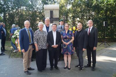 Eine Gruppe von vier Männern und zwei Frauen stehen in Zweierreihen vor einem Steingrabmal und schauen in die Kamera. Im Hintergrund Menschen, Bäume und Blumenkränze, die um die Grabstätte drapiert sind. 