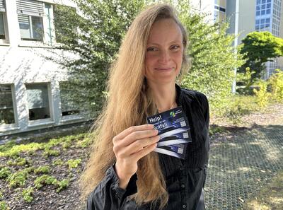 Eine Frau Ende 30 mit langen, offenen dunkelblonden Haaren hält lächelnd drei Karten mit der englischen Aufschrift "Help" in der rechten Hand und lächelt in die Kamera. 