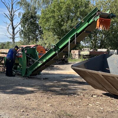 Ein Mann im Blaumann mit Schallschutzkopfhörern bedient eine landwirtschaftliche Anlage, in der Baumstämme zu Kamin- und Brennholz verarbeitet werden.