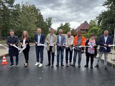 Männer und Frauen stehen mit Scheren in den Händen in einer Reihe auf einer Straße und durchschneiden ein weißes Band. 