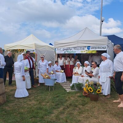 Weiße Zelte mit polnisch-beschrifteten Schildern stehen auf einer Wiese. Im Vordergrund Frauen und Männer in weißen Trachten.