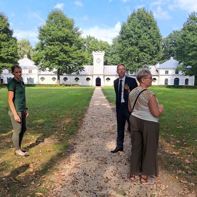 Zwei Frauen und ein Mann stehen auf einem Weg. Links und rechts Wiese und Bäume. Im Hintergrund ein Gutsschloss und Pferdegestüt.