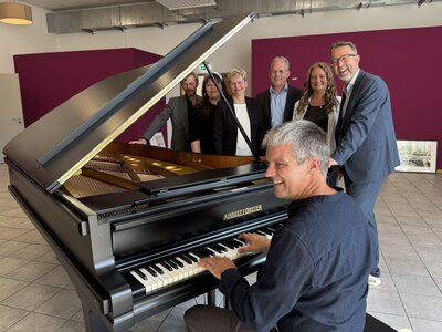 Ein Mann mit grauen Haaren und langärmeligem Hemd spielt am Klavier. Drei Männer und drei Frauen in festlicher Kleidung stehen hinter dem Klavier und lächeln in die Kamera.