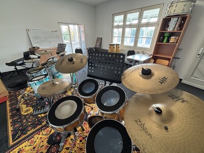 Eine Vielzahl an Schlagzeugen und ein Notenständer stehen in einem hellen Raum mit Regalen und bodentiefem Fenster. 