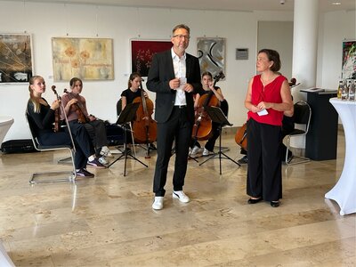 Ein Mann im Anzug und eine Frau mit roter Bluse stehen vor einer Musikerinnengruppe. 