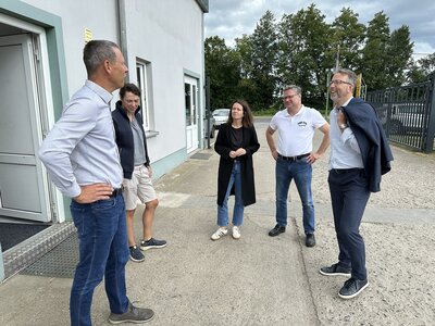 Drei Männer und zwei Frauen stehen in einem Halbkreis zusammen und unterhalten sich. Rechts im Bild ein weißes Haus mit Fenstern und einer geöffneten Eingangstür. 