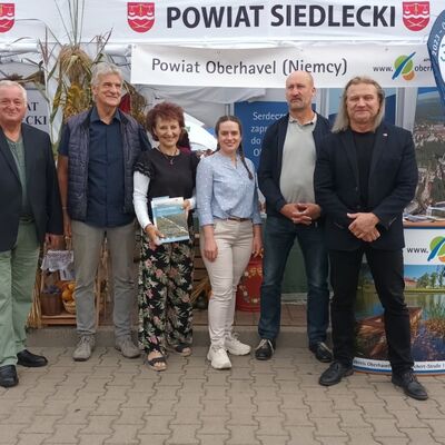 Eine Gruppe von vier Männern und zwei Frauen steht vor einem Messestand, auf dem mit polnischer Schrift Powiat Siedlecki und Powiat Oberhavel (Niemcy) steht. Die Personen schauen frontal in die Kamera.