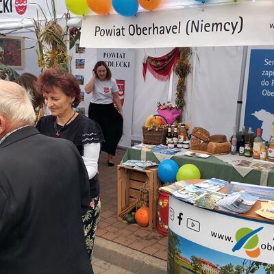 Männer und Frauen stehen links im Bildvordergrund. Eine Frau mit einem schwarzem Rock im Bildhintergrund steht neben dem gedeckten Tisch des Messestandes, auf dem es typische Köstlichkeiten wie Brot, Säfte gibt. Oberhalb des Tisches hängt ein auf polnisch bedrucktes Banner auf dem steht: Powiat Oberhavel (Niemcy).