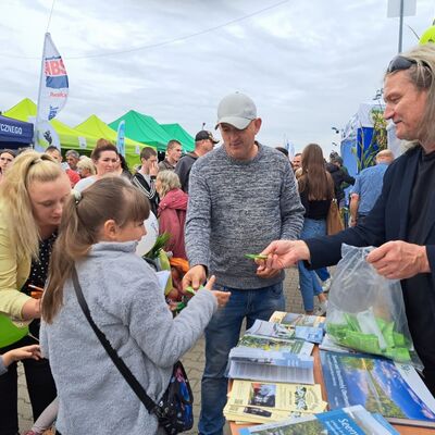 Ein Mann in schwarzem Shirt und mit schwarzem Sakko hält in der linken Hand eine durchsichtige Tüte mit Gummibärchen in grünen Plastiktütchen. Ein Tütchen überreicht er einem Mädchen im grauen Pulli mit langen, dunkelblonden Haaren mit Pony, die zu einem Pferdeschwanz zusammengebunden sind.