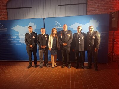 Fünf Männer in dunklen Uniformen und eine Frau mittleren Alters stehen in einer Reihe vor einer Aufstellerwand des Landes Brandenburgs und schauen in die Kamera. 