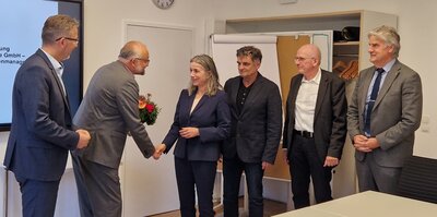 Ein älterer Herr im grauen Anzug reicht einer Frau mittleren Alters im blauen Anzug die Hand. Drumherum stehen im Halbkreis vier weitere Herren in Anzügen in einem Büro. 