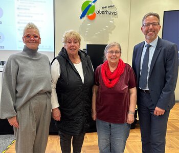 Eine Gruppe von vier Personen, davon drei ältere Frauen und ein Mann im blauen Anzug mittleren Alters. 