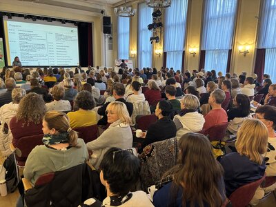 Eine große Gruppe an Menschen, sowohl Frauen als Männer, sitzen auf Stühlen in mehreren Reihen in einem großen Saal und schauen auf eine Beamerleinwand nach vorne zu einer Bühne. 
