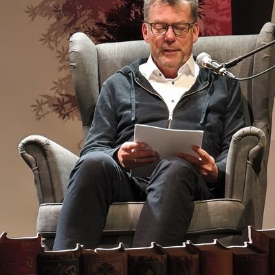 Autor Richard Brandes sitzt in einem Sessel auf der Bühne und liest aus seinem Krimi "Gejagt durch Brandenburg"