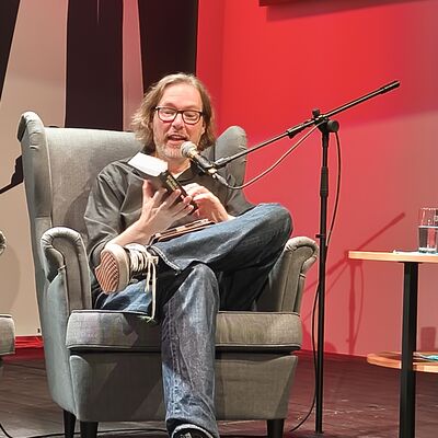 Der Autor Sven Stricker sitzt in einem Sessel auf der Bühne und liest aus seinem neusten Buch.