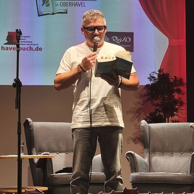 Der Autor Andreas Winkelmann steht auf der Bühne und liest aus seinem humorvollen Campingkrimi "Entführung im Himmelreich".