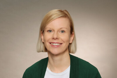 Eine Frau mittleren Alters mit blonden, kinnlangen Haaren, grünen Augen und grüner Jacke und weißem Shirt. 