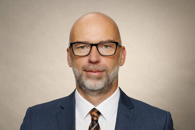 Ein Mann mit schwarzer Brille mittleren Alters ohne Haare, graumeliertem Vollbart mit dunkelblauem Sakko, dunkelgemusterter Krawatte und weiem Hemd.