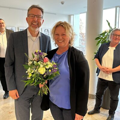 Im Vordergrund: Ein Mann im grauen Anzug und weißem Hemd. Neben ihm eine Frau mit blonden, kurzen Locken und blauem Shirt und schwarzem Oberteil.