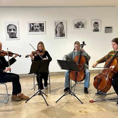 Vier Mädchen sitzen auf Stühlen und spielen Musikinstrumente. Zwei spielen Geige, zwei Mädchen Kontrabass.